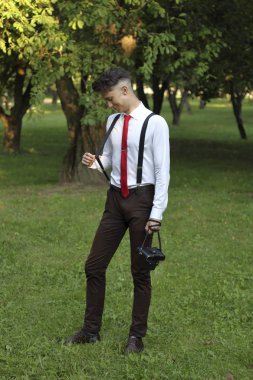 Şık giyimli genç adam bir parkta poz. Sakızı idrardan çıkarır. Bir Slr film kamera omzuna asılı.