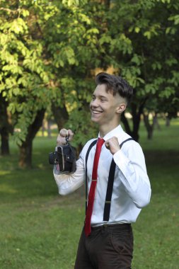 Şık giyimli genç adam bir parkta poz. Kamerayı elinde tutuyor ve gülüyor, yumruklarını sıkıyor..