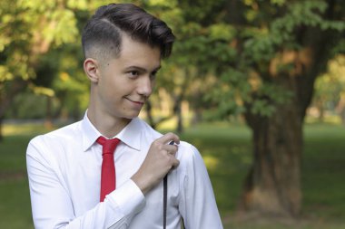 Şık giyimli genç adam bir parkta poz. Kameradaki kayışı düzeltir.