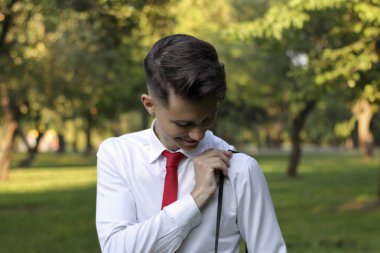 Şık giyimli genç adam bir parkta poz. Kameranın omzundaki kayışı güler ve ayarlar.