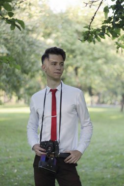 Şık giyimli genç adam bir parkta poz. Bir Slr film kamera boyun asılı.