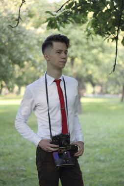 Şık giyimli genç adam bir parkta poz. Bir Slr film kamera boyun asılı.