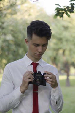 Şık giyimli genç adam bir parkta poz. Kamerayı elinde tutar ve ayarlarla çalışır.
