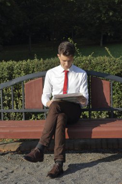 Şık giyimli genç adam bir park bankına oturur ve bir gazete okur.