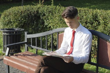 Şık giyimli genç adam bir park bankına oturur ve bir gazete okur.