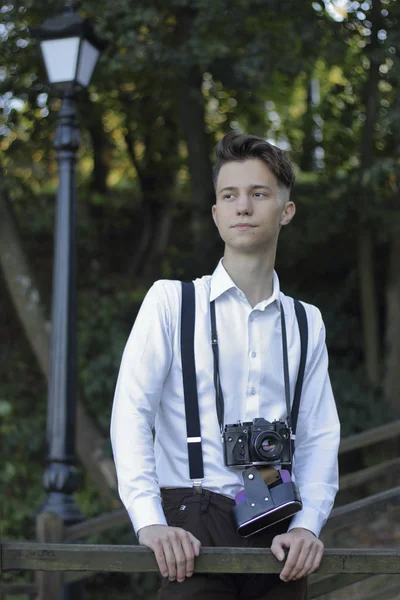 Şık giyimli genç adam bir parkta poz. Bir Slr film kamera boyun asılı.