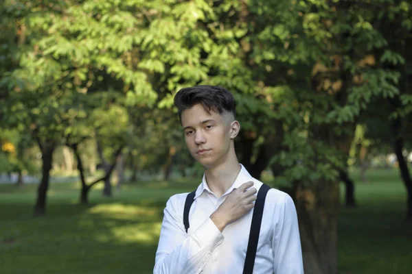 Stylishly dressed young man posing in a park. Looks thoughtfully to the side, straightens his shirt collar. — Stock Photo, Image