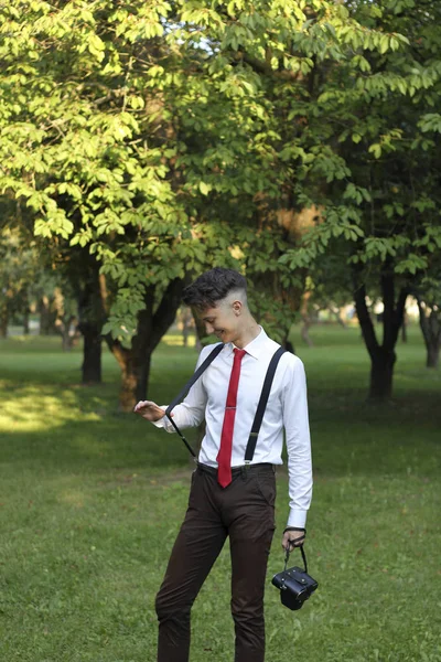 Şık giyimli genç adam bir parkta poz. Sakızı idrardan çıkarır. Bir Slr film kamera omzuna asılı. — Stok fotoğraf