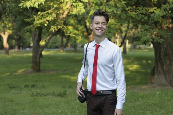 Jeune homme élégamment habillé posant dans un parc. Un appareil photo reflex est accroché à son épaule . — Photo