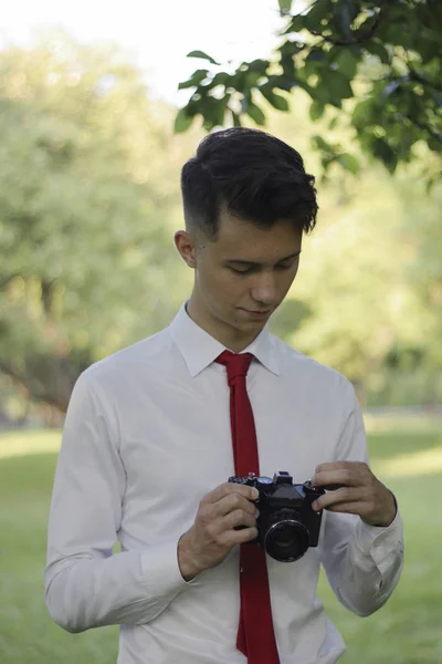 Стильно одетый молодой человек позирует в парке. Держит камеру в руках и работает с настройками . — стоковое фото