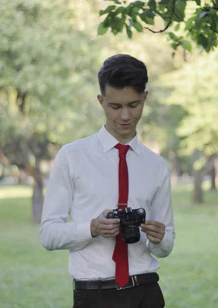 Stylowo ubrany młody mężczyzna stwarzając w parku. Trzyma aparat w dłoniach i współpracuje z ustawieniami. — Zdjęcie stockowe
