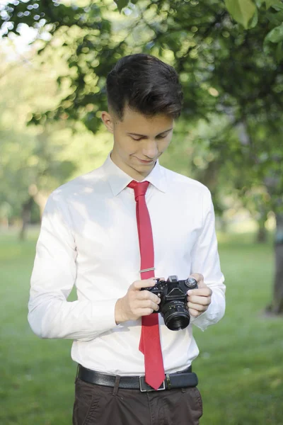 Şık giyimli genç adam bir parkta poz. Kamerayı elinde tutar ve ayarlarla çalışır. — Stok fotoğraf