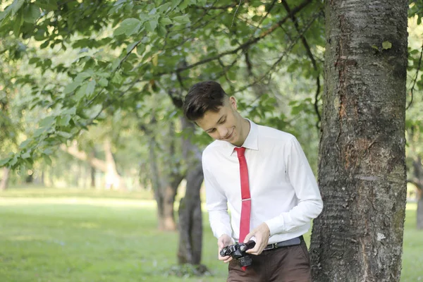 Jovem elegantemente vestido posando em um parque. Segura a câmera em suas mãos e trabalha com as configurações . — Fotografia de Stock