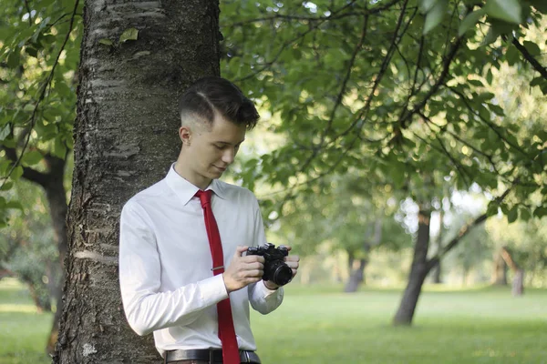 Stijlvol gekleed jonge man poseren in een park. Houdt de camera in zijn handen en werkt met de instellingen. — Stockfoto