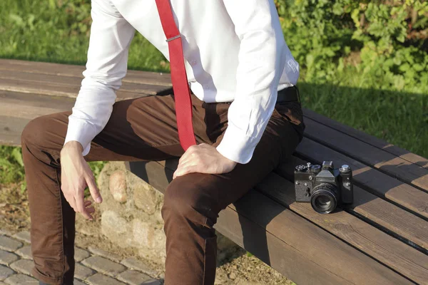 Élégamment habillé jeune homme assis sur un banc de parc. A proximité se trouve un appareil photo reflex . — Photo