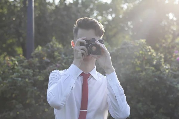 Le jeune homme élégamment habillé est assis en train de prendre des photos dans le parc. Dans ses mains tient un appareil photo reflex . — Photo