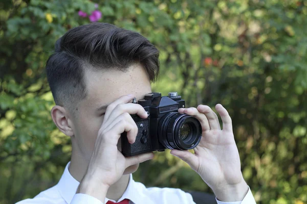 Le jeune homme élégamment habillé est assis en train de prendre des photos dans le parc. Dans ses mains tient un appareil photo reflex . — Photo