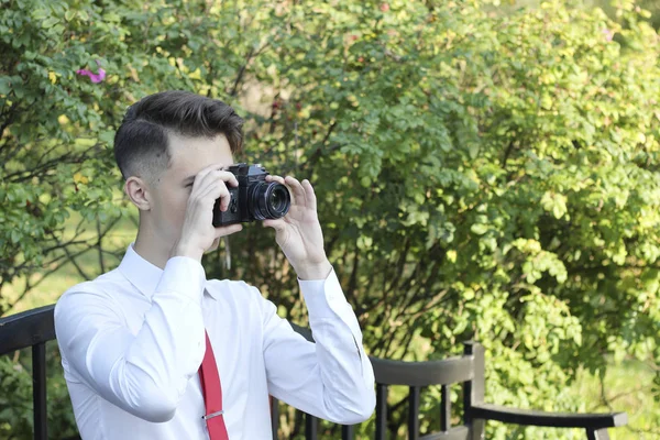 Giovane uomo elegantemente vestito si siede a scattare foto nel parco. Nelle sue mani tiene una fotocamera reflex . — Foto Stock