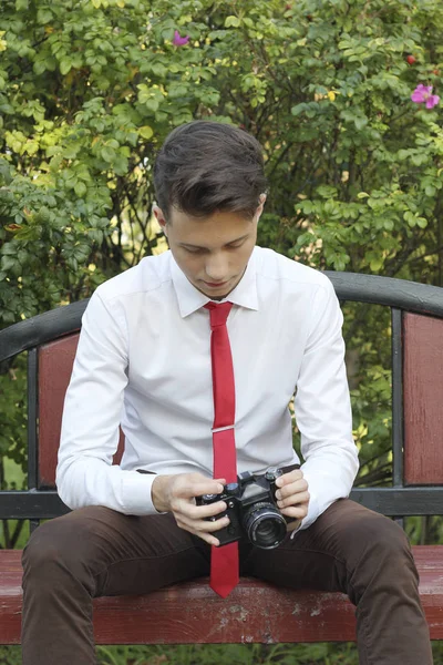Un jeune homme élégamment habillé s'assoit sur un banc de parc. Élégamment habillé jeune homme assis sur un banc de parc. Dans ses mains tient un appareil photo reflex . — Photo