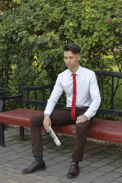 Stylowo ubrany młody człowiek siedzi na ławce w parku i trzyma gazetę w dłoniach. spogląda z dala z uśmiechem. — Zdjęcie stockowe