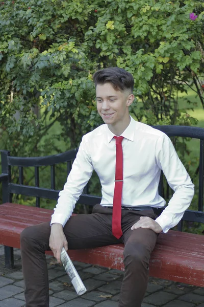 Un joven vestido con estilo se sienta en un banco del parque y tiene un periódico en sus manos. mira hacia otro lado con una sonrisa . —  Fotos de Stock