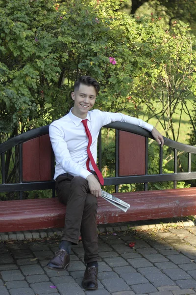 Stylowo ubrany młody człowiek siedzi na ławce w parku i trzyma gazetę w dłoniach. spogląda z dala z uśmiechem. — Zdjęcie stockowe