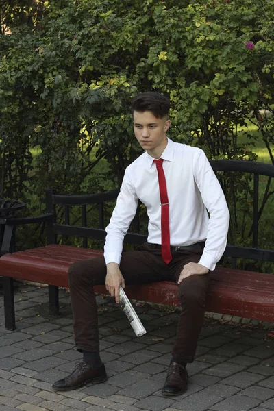 Un jeune homme élégamment habillé s'assoit sur un banc de parc et tient un journal entre ses mains. regarde loin avec un sourire . — Photo