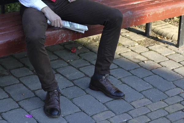 Un jeune homme élégamment habillé s'assoit sur un banc de parc et tient un journal entre ses mains. Fermer les jambes visibles . — Photo