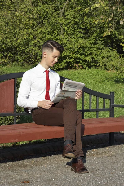 Jeune homme élégamment habillé assis sur un banc de parc et lit un journal . — Photo