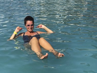 Açık havada havuzdaki kız. Havuzun tuzlu suyu onu su yüzeyinde tutar. Gülümseyen, başında güneş gözlüğü.