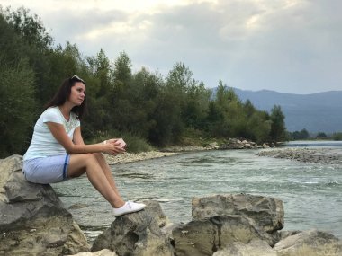 A girl sits on a large boulder near a mountain river. Holds a telephone in his hands and looks at the water stream.