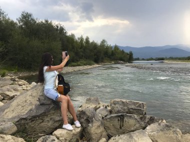 A girl sits on a large boulder near a mountain river. Near a tourist backpack. She looks at the water stream and takes pictures.