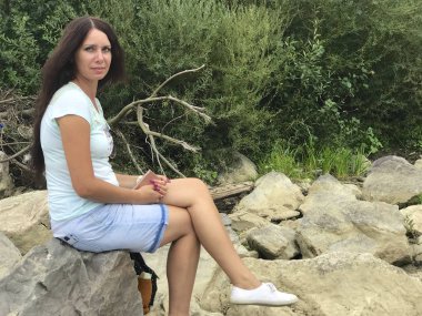 A girl sits on a large boulder near a mountain river. Near a tourist backpack. She is looking into the camera lens.