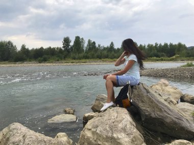 A girl sits on a large boulder near a mountain river. backpack. She is looking at the water stream.
