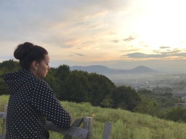 Bir kız yaylalarda bir yamaç üzerinde ahşap bir çit duruyor. Gün batımında mesafeye bakar.
