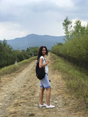 Omzunda sırt çantası olan bir kız bir köy yolunda ve etrafında dönüyor. Ufukta çok uzakta sisle kaplı dağlar var..