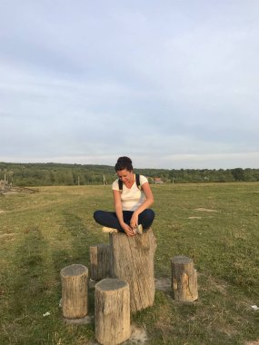 Arkasında sırt çantası olan bir kız. Tahta bir kütüğün üzerinde oturur. Yaylalar arasında bir vadide.