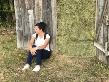 Arkasında sırt çantası olan bir kız. Saman dolu ahşap bir baraka tarafından oturur.