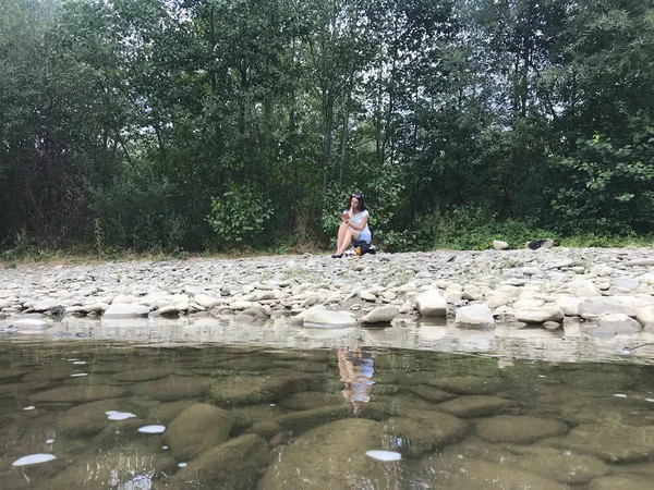 Девушка сидит на камне на берегу горной реки. Посмотри в его телефон. Поток воды медленно течет мимо . — стоковое фото