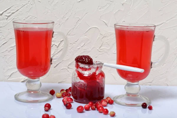 Zelfgemaakte blanks. Cranberry jam in een open pot en glazen met cranberry sap. Op een schotel is een theelepel met jam. Verschillende bessen zijn verspreid over het oppervlak. — Stockfoto