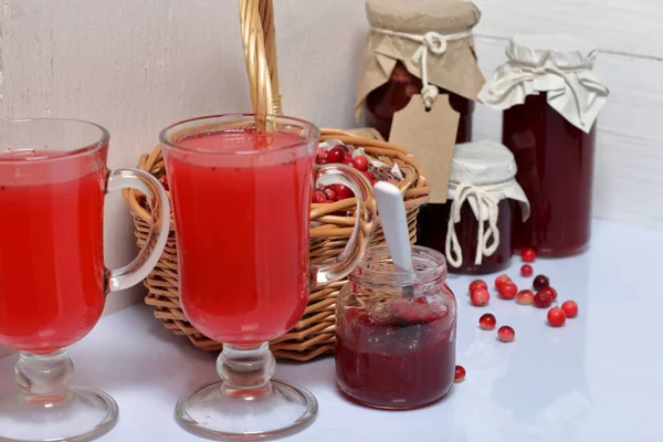 Homemade blanks. Cranberry jam in jars and glasses with cranberry juice. One can is open, it has a teaspoon. Several berries are scattered on the surface.