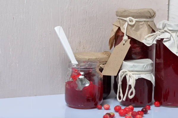 Homemade preserves, cranberry jam in jars. One can is open, it has a teaspoon. Several berries are scattered on the surface.