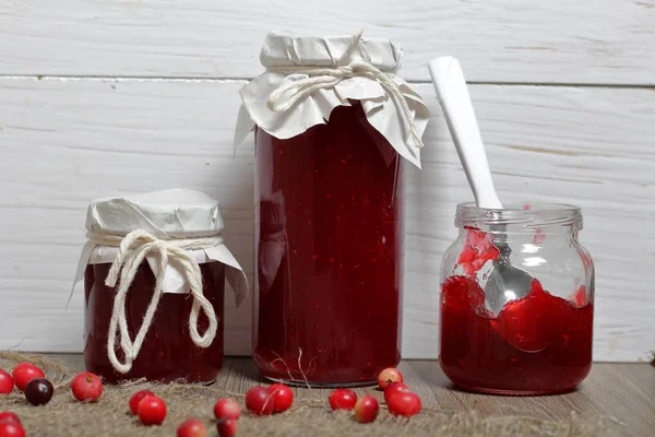 Homemade preserves, cranberry jam in jars. One can is open, it has a teaspoon. Several berries are scattered on the surface.
