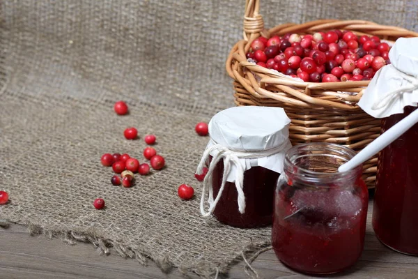 Homemade preserves, cranberry jam in jars. One can is open, it has a teaspoon. Several berries are scattered on the surface.