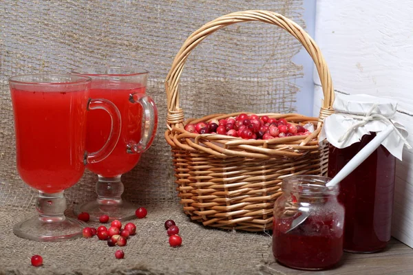 Homemade blanks. Cranberry jam in jars and glasses with cranberry juice. One can is open, it has a teaspoon. Several berries are scattered on the surface.