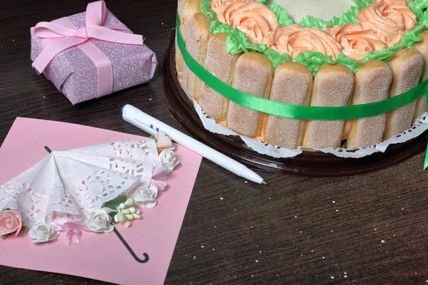 Pastel de esponja con queso crema, decorado con galletas Savoiardi. En el lateral, envuelto en una cinta atada a un lazo. Muy cerca hay una tarjeta de felicitación, regalo y pluma . — Foto de Stock