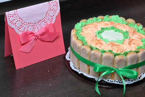 Gâteau éponge au fromage à la crème, décoré de biscuits Savoiardi. Sur le côté, enveloppé dans un ruban attaché à un arc. Près d'une carte de voeux . — Photo