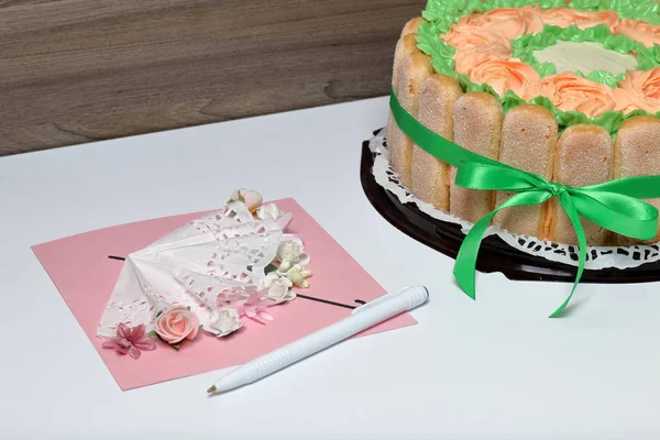 Pastel de esponja con queso crema, decorado con galletas Savoiardi. En el lateral, envuelto en una cinta atada a un lazo. Cerca de una tarjeta de felicitación y pluma . — Foto de Stock