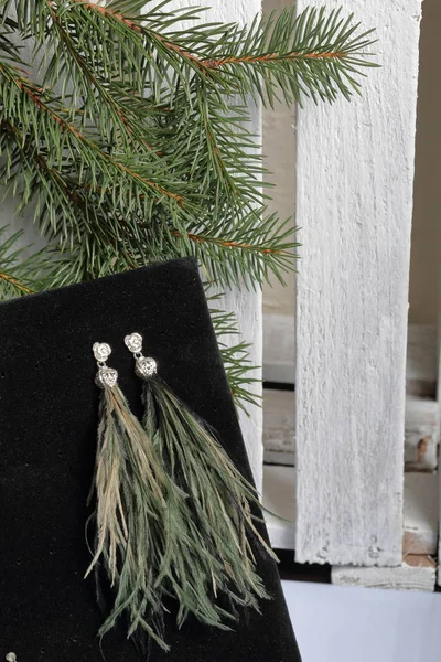 Ostrich feather brush earrings. On black velvet. Against the background of a fir branch and a white wooden box.