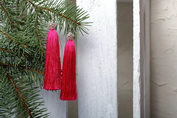 Pink tassel earrings hang on a spruce branch. Against the background of a white wooden box.